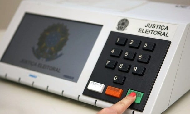 Modelo de urna eletrônica usada no sistema eleitoral brasileiro. Na imagem, mostra-se um dedo indicador direito, possivelmente de uma mulher, pois está com esmalte vermelho na unha, em que simula-se o momento final da votação: teclando o botão verde, de 'confirma'